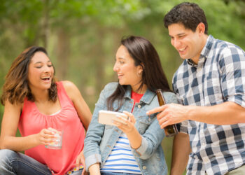 Two young women and one man view a social media post on their cell phone. The woman in center is holding the smart phone while the two friends  view the post, text message, tv show, or video.  Latin descent, caucasian, and mixed race group of people. Beautiful spring or summer nature background. Park, or backyard setting.
