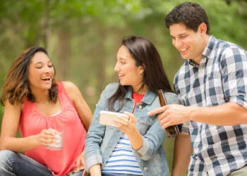 Two young women and one man view a social media post on their cell phone. The woman in center is holding the smart phone while the two friends view the post, text message, tv show, or video. Latin descent, caucasian, and mixed race group of people. Beautiful spring or summer nature background. Park, or backyard setting.