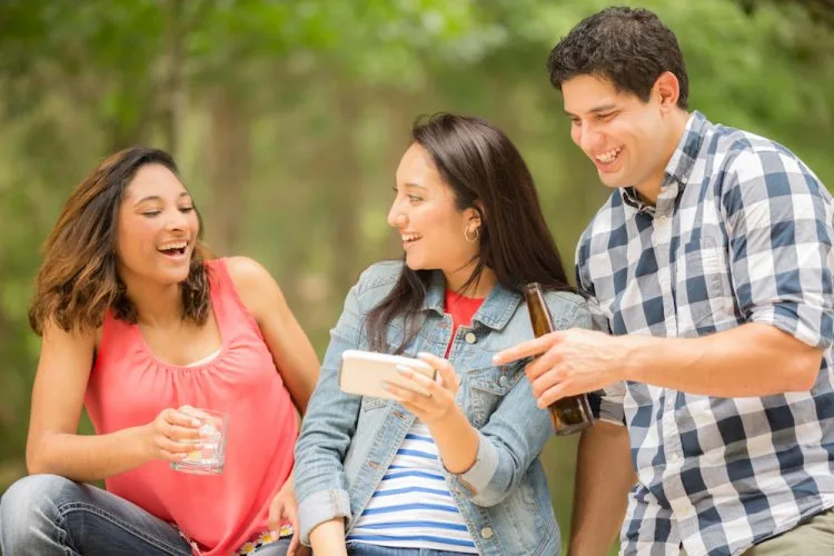 Two young women and one man view a social media post on their cell phone. The woman in center is holding the smart phone while the two friends view the post, text message, tv show, or video. Latin descent, caucasian, and mixed race group of people. Beautiful spring or summer nature background. Park, or backyard setting.